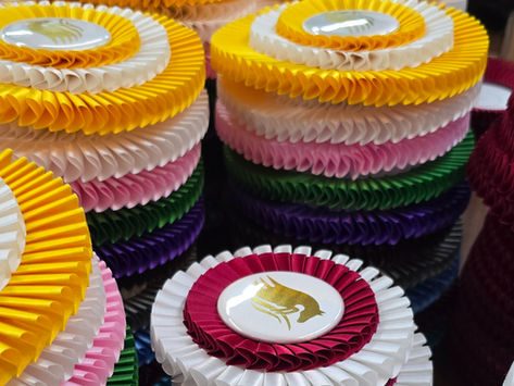 Stacks of colorful rosette award ribbons in yellow, white, pink, green, purple, and red, neatly arranged on a table.