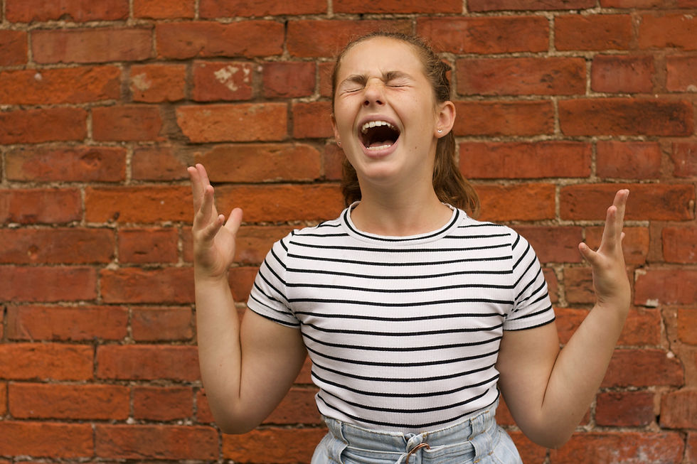 Teen girl in striped shirt screams or sings with eyes shut and hands raised against a red brick wall.