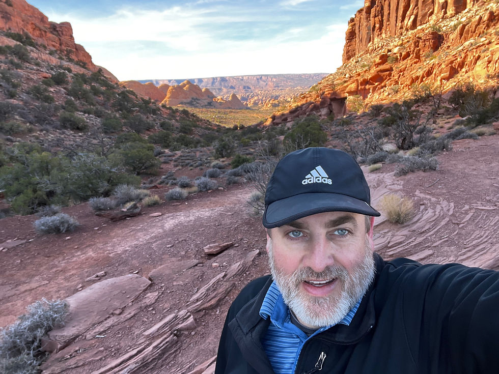 Man in a black Adidas cap smiles, standing on a rocky path in a canyon. Red cliffs and a vast vista are in the sunlit background.