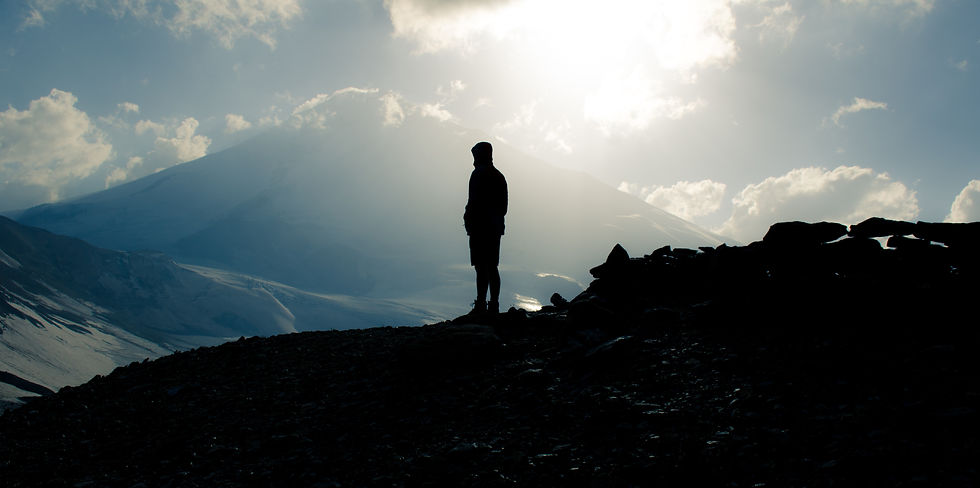 Silhouette of a person standing on a rocky mountain ridge beneath bright sun and clouds, overlooking snowy peaks