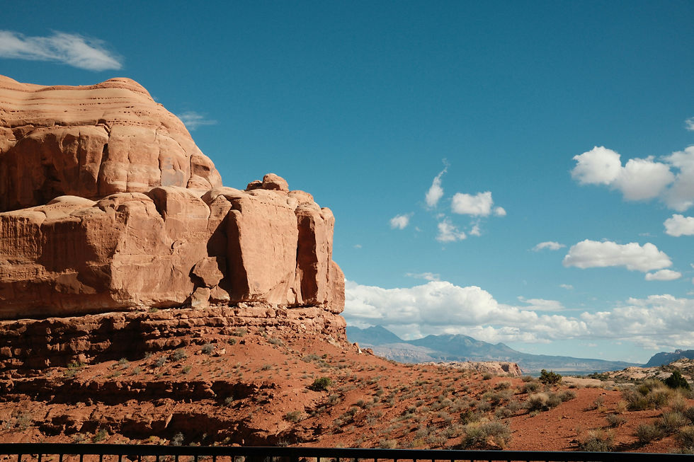 Red rock formations under a bright blue sky with scattered clouds, desert landscape, and distant mountains in the background. Calm and scenic.