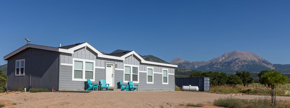 Gray house with turquoise chairs in desert setting, mountains in the background, under clear blue sky. Tranquil and isolated atmosphere.