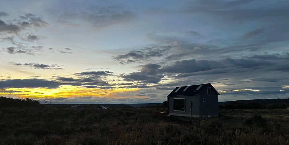 A small gray house sits in a field under a dramatic sunset with clouds. The sky has hues of yellow and blue, creating a serene mood.