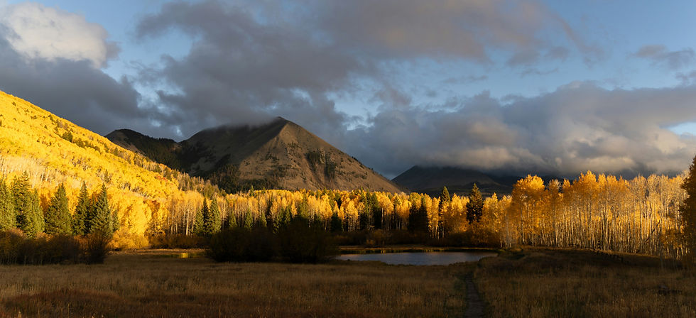 Golden fall trees surround a serene pond at the base of mountains under a partly cloudy sky, creating a peaceful autumn landscape.