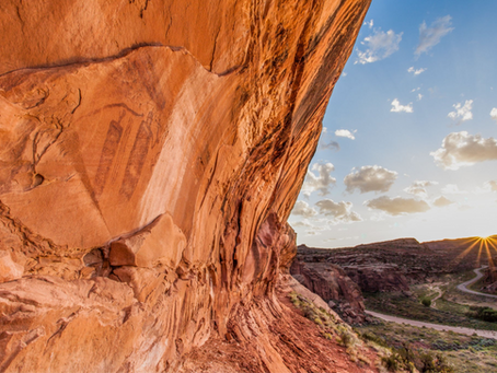 Massive orange sandstone cliff with petroglyphs over a winding canyon road at sunset, under a blue sky with clouds.