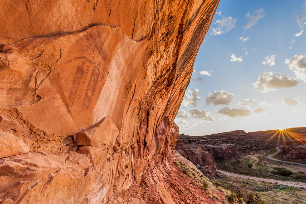 Massive orange sandstone cliff with petroglyphs over a winding canyon road at sunset, under a blue sky with clouds.