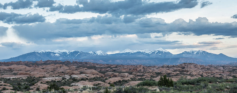 Rocky desert foreground with snow-capped mountains under a dramatic cloudy sky, wide panoramic landscape.