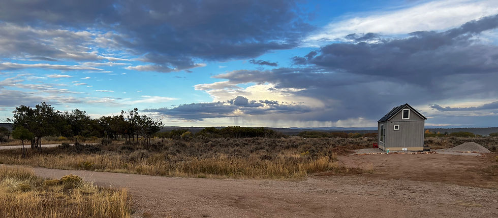 Small gray tiny house in a dry field under dramatic storm clouds and blue sky, with distant rain over open scrubland.