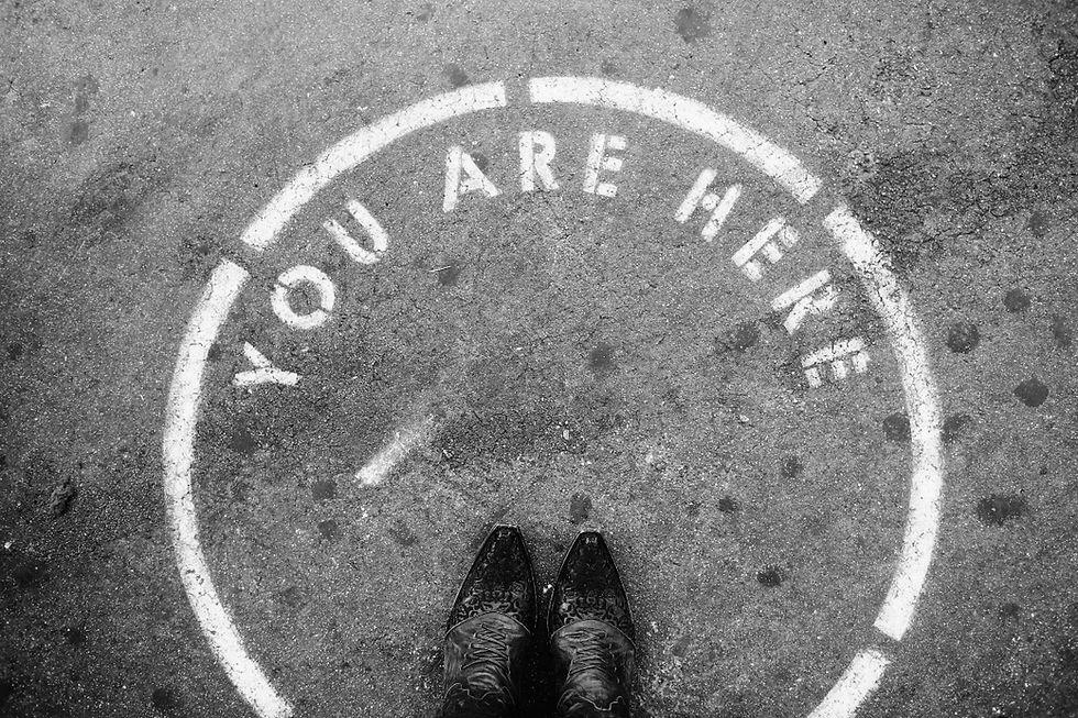 Black-and-white pavement with a painted YOU ARE HERE circle; polished boots stand at the bottom, giving a reflective mood.