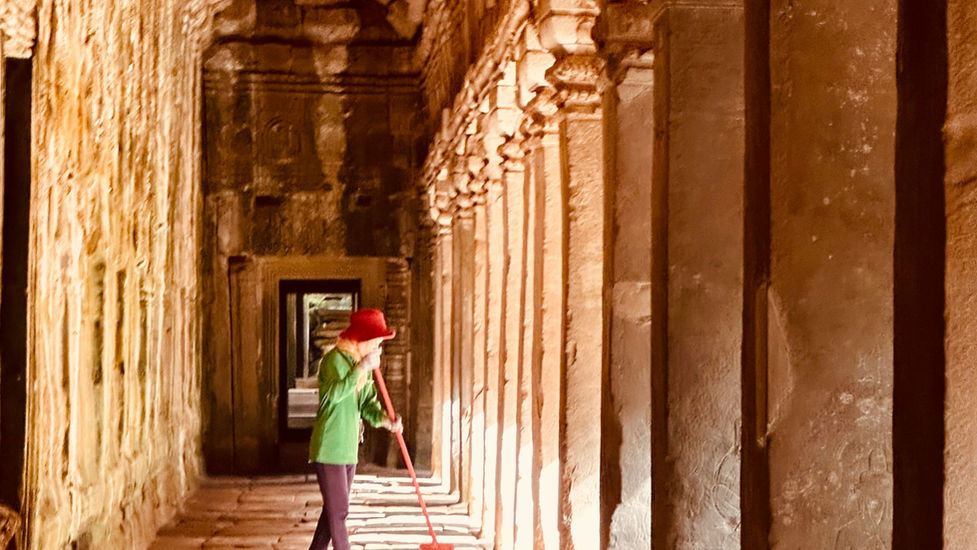 Person in red hat and green shirt sweeping inside an ancient stone corridor, warm light highlighting intricate carvings and pillars.