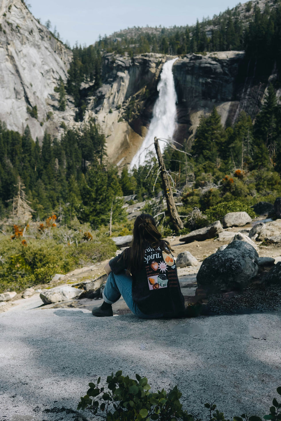 Person sits on a rocky overlook facing a tall waterfall in a pine forest, wearing a black graphic T-shirt.