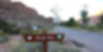 Sign for "Moab Rim" on a dirt road, with red rock cliffs and greenery in the background under a clear sky.