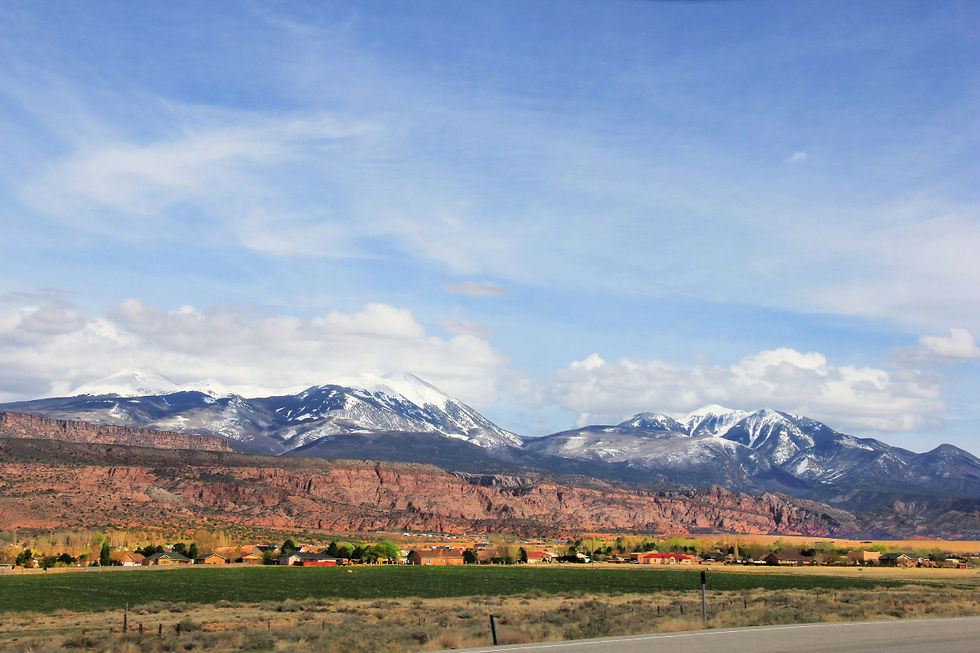 Snow-capped mountains under a blue sky with scattered clouds. Red rock formations and green fields with houses are visible in the foreground.