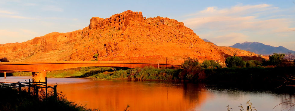 Sunset over orange desert mesa with a bridge crossing a calm river, reflecting warm light and dark shrubs.
