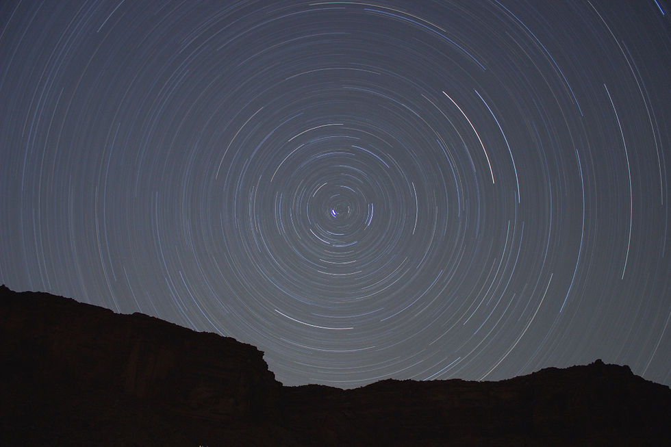 Circular star trails swirl across a dark night sky above silhouetted mountains, creating a calm, cosmic scene.