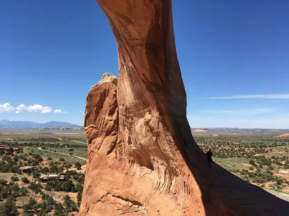A look from under the arch, the vast landscape looking west toward Monticello, UT. It has green fields and random green trees with mountains in the backdrop to the left. There are a few houses at the bottom-left of the picture.
