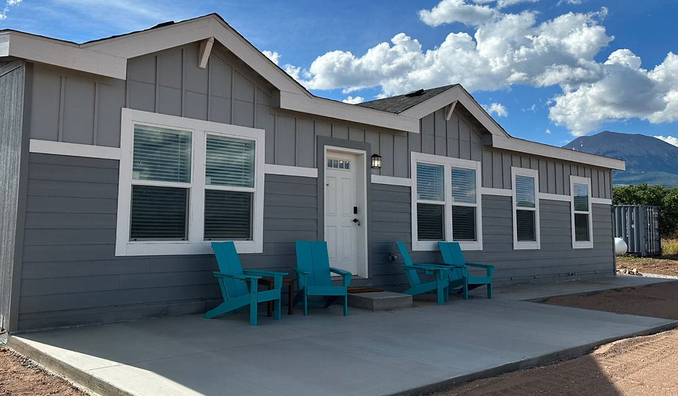 Gray house with white trim, adorned with teal Adirondack chairs on the porch. Blue sky with clouds and mountains in the background.