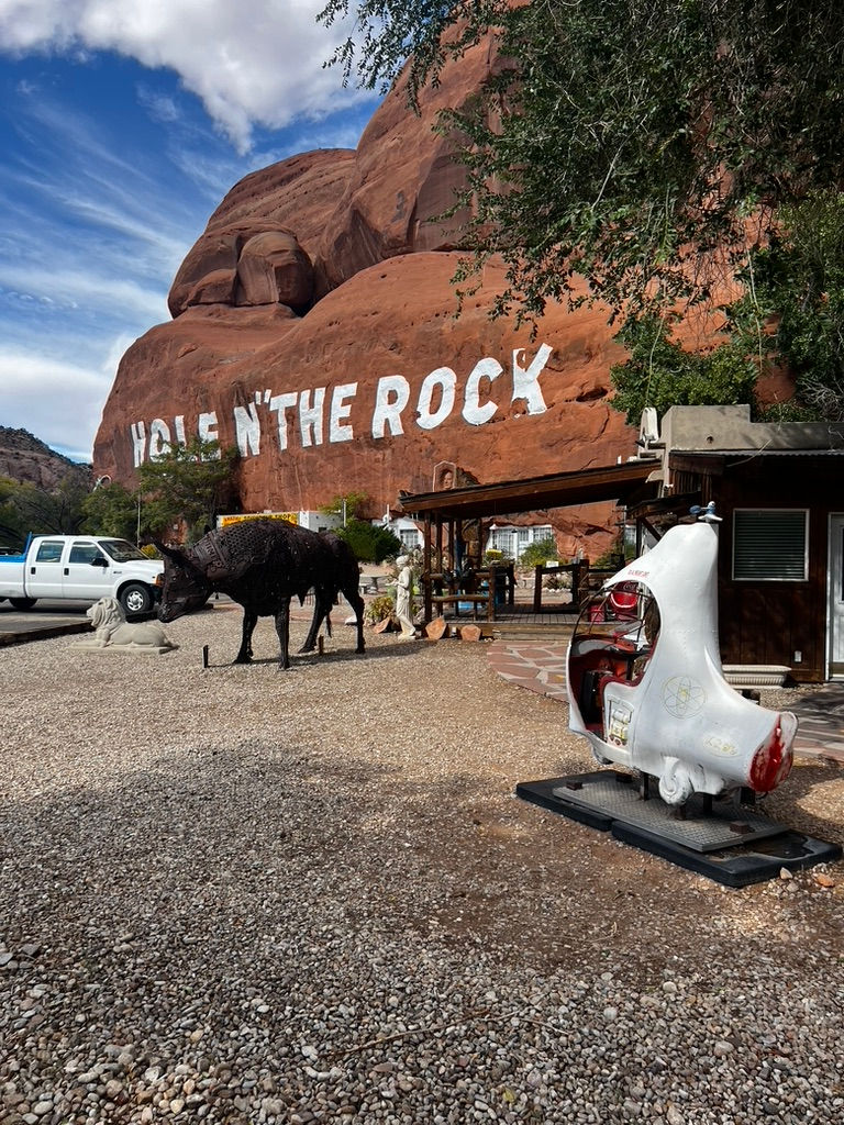Red rock formation with "Hole N' The Rock" text, a buffalo sculpture, and a white ride-on aircraft. Blue sky, trees, and a white truck.