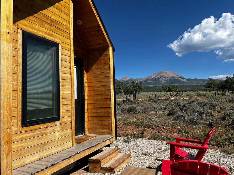Wooden cabin with red chairs overlooks a dry field and mountain under a bright blue sky with a large white cloud.