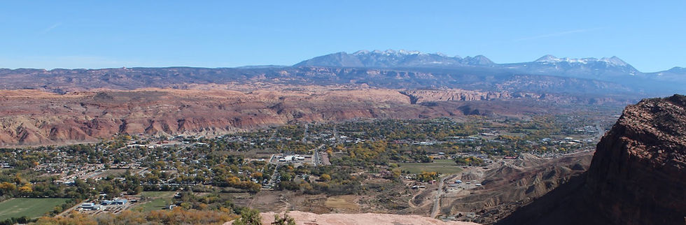 Panoramic valley town below red cliffs and snowcapped mountains under a clear blue sky, with a calm, expansive feel