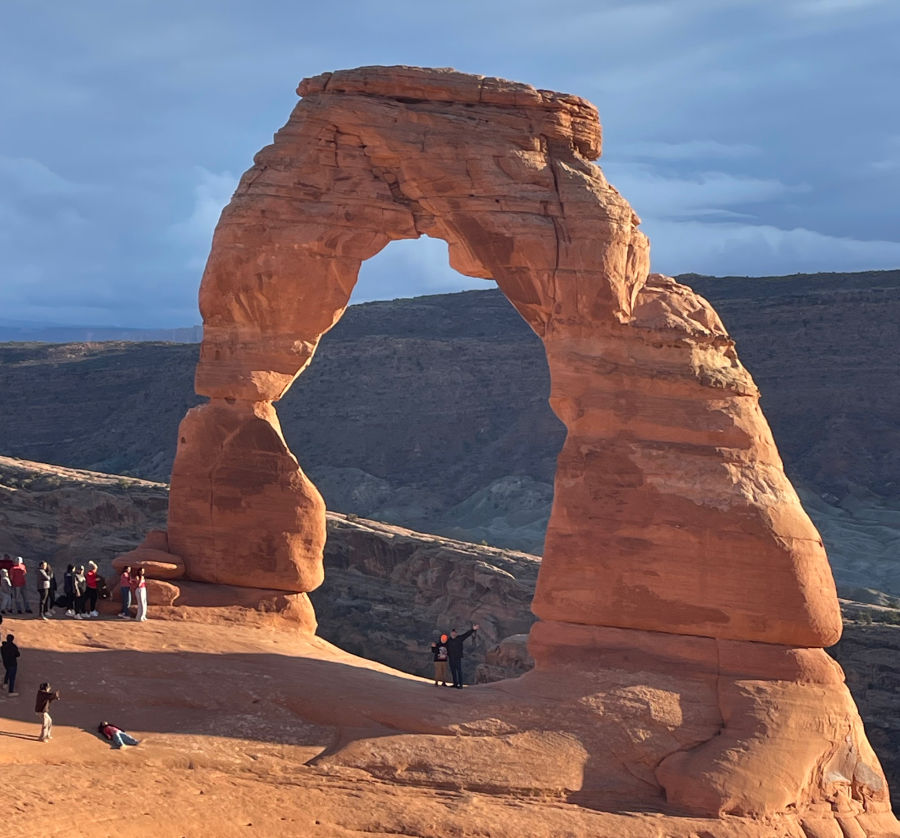 Sunlit red sandstone Delicate Arch in a canyon, with a few hikers posing and taking photos under a cloudy blue sky.