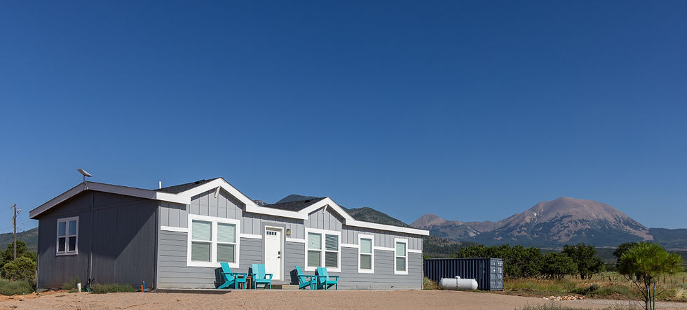 Gray house with turquoise chairs on a dirt lot, backed by mountains under a clear blue sky, calm rural scene.