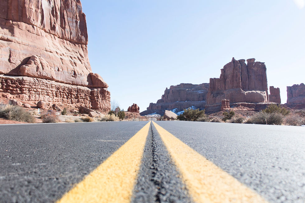 Desert road with yellow lines leads to towering red rock formations under a clear blue sky. Sparse vegetation and rugged terrain surround.