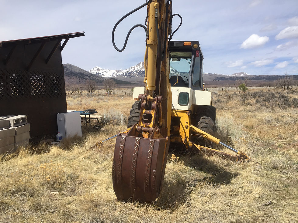 Yellow backhoe in a grassy field, mountains in background, near a dark wooden shed with cinder blocks. Cloudy sky and rugged terrain.