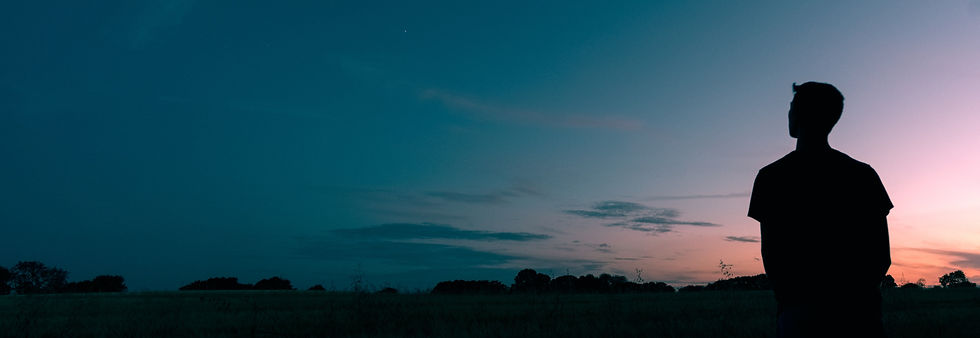 Silhouette of a person standing in a field at sunset. Sky is a gradient of blue to pink with scattered clouds, creating a calm atmosphere.