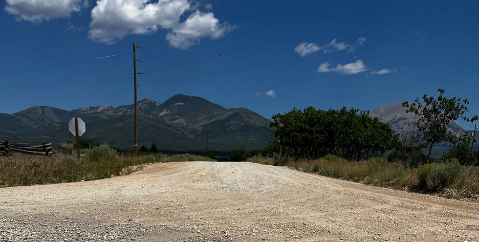 Gravel road leading toward mountains under a bright blue sky, with a stop sign, power pole, and trees on a quiet rural scene.