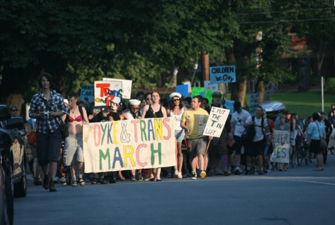 A group of people participating in a "Dyke & Trans March" on a tree-lined street. Participants hold a large hand-painted banner and various signs