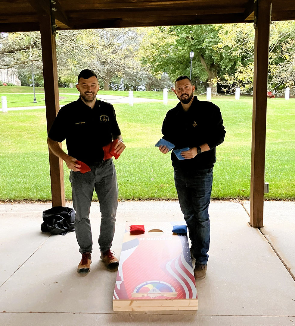 Two lodge brothers in black lodge shirts holding cornhole bags beside a board under a pavilion.