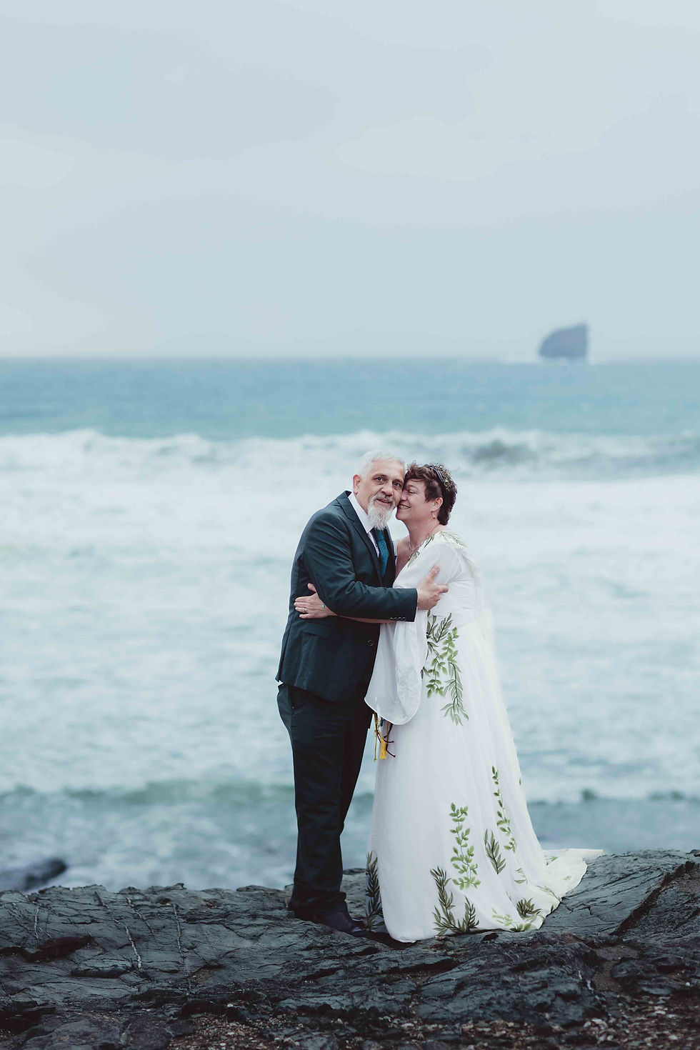 An elderly couple embraces on a rocky shore, the woman in a white dress with green patterns. Ocean waves and a misty sky set a serene mood.