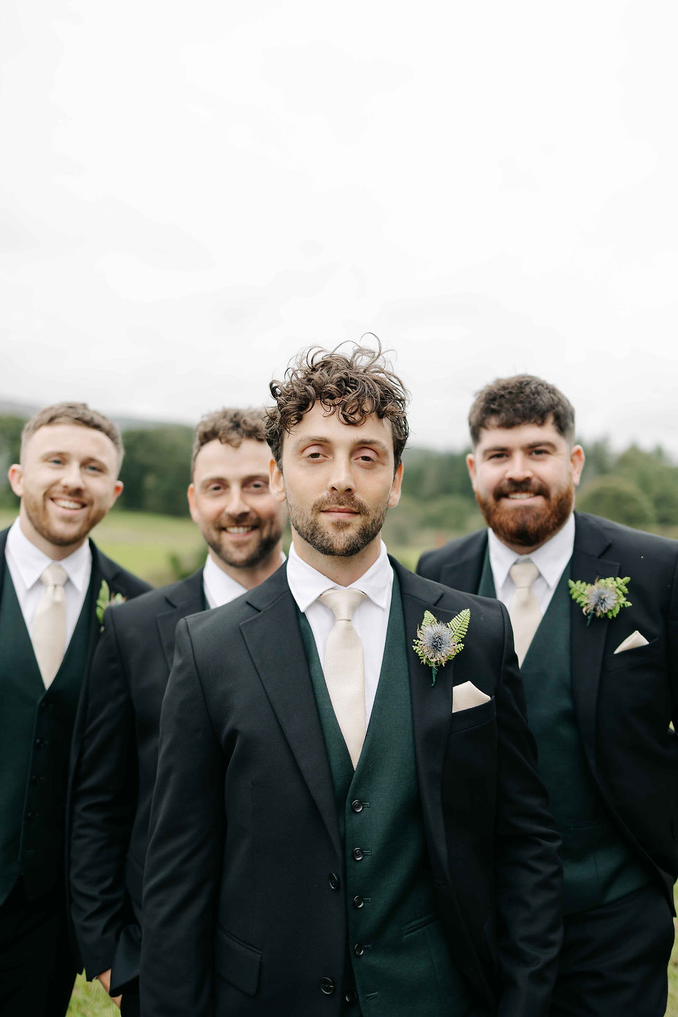 Four groomsmen in black suits and cream ties pose smiling outdoors with green fields behind them.