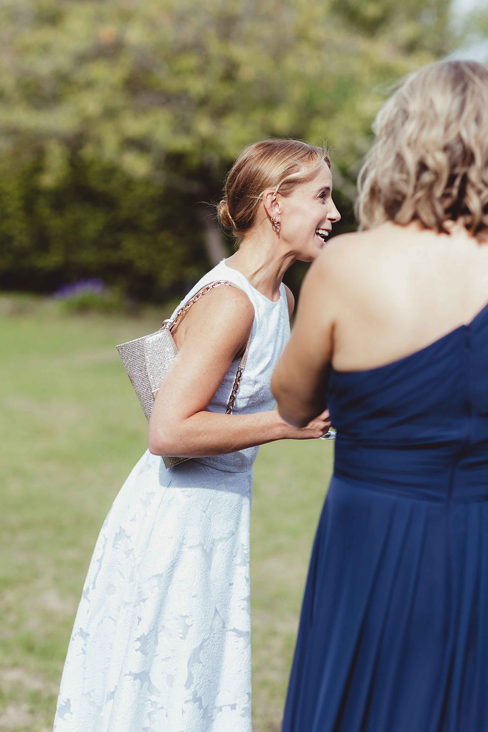 Two women chatting and laughing outdoors. One wears a white dress and carries a shiny purse. The other wears a navy dress. Greenery behind.