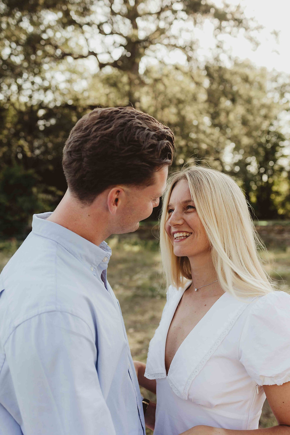 A couple smiles at each other in a sunny park setting. The woman wears a white dress, the man a light blue shirt. Greenery in the background.