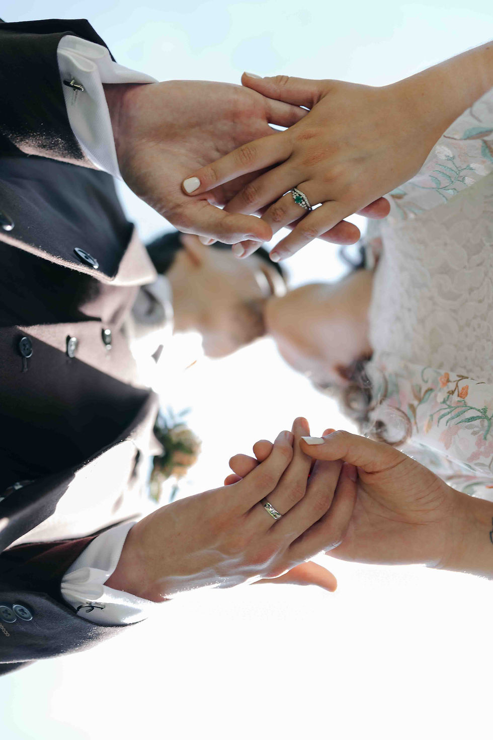 Bride and groom clasp hands, showing wedding rings, with lace dress and dark suit against a bright sky.