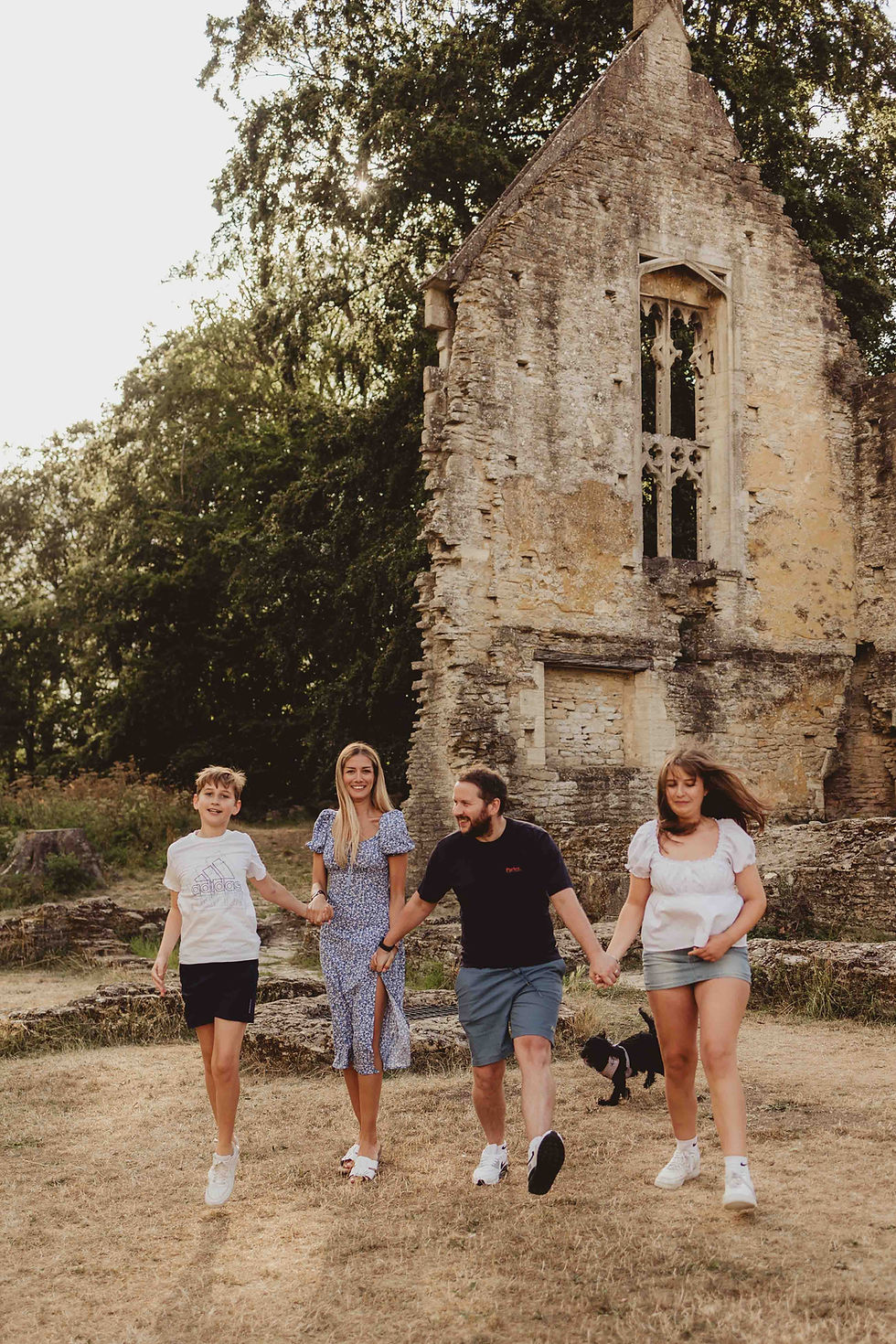 A family of four and a small dog joyfully walk hand in hand by a stone ruin, set in a sunlit, grassy area with trees in the background.