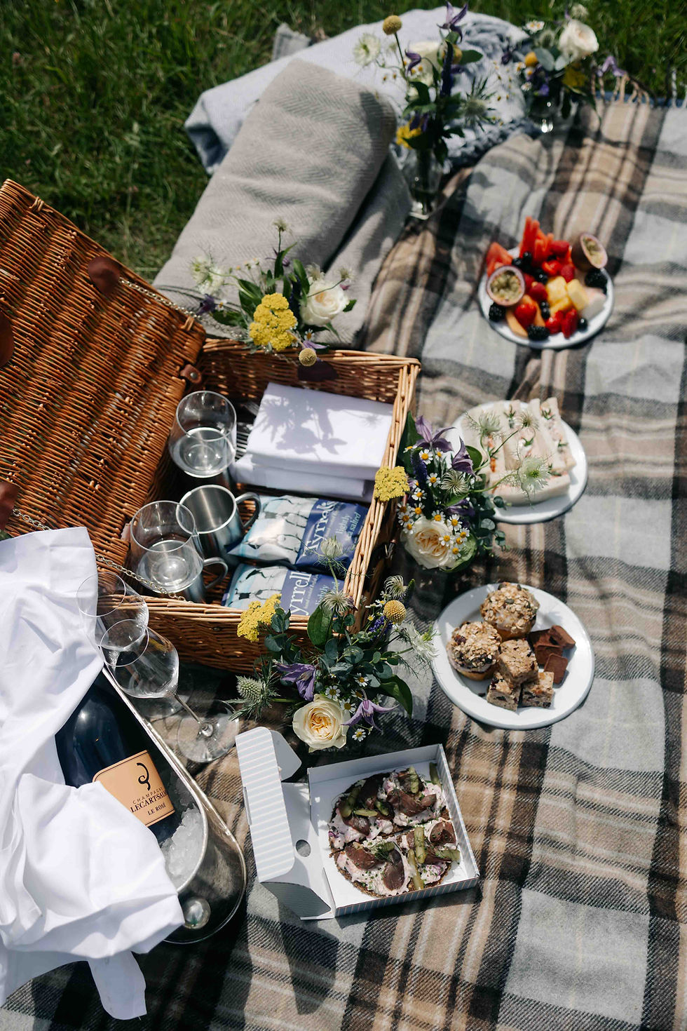 Elegant picnic on a plaid blanket with wicker basket, flowers, champagne, fruit and desserts in a sunny grassy field.