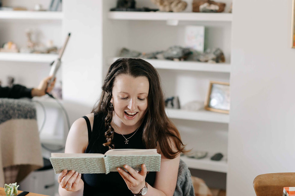 Smiling woman reads a book in a cozy room with white shelves and decor, looking delighted and absorbed.