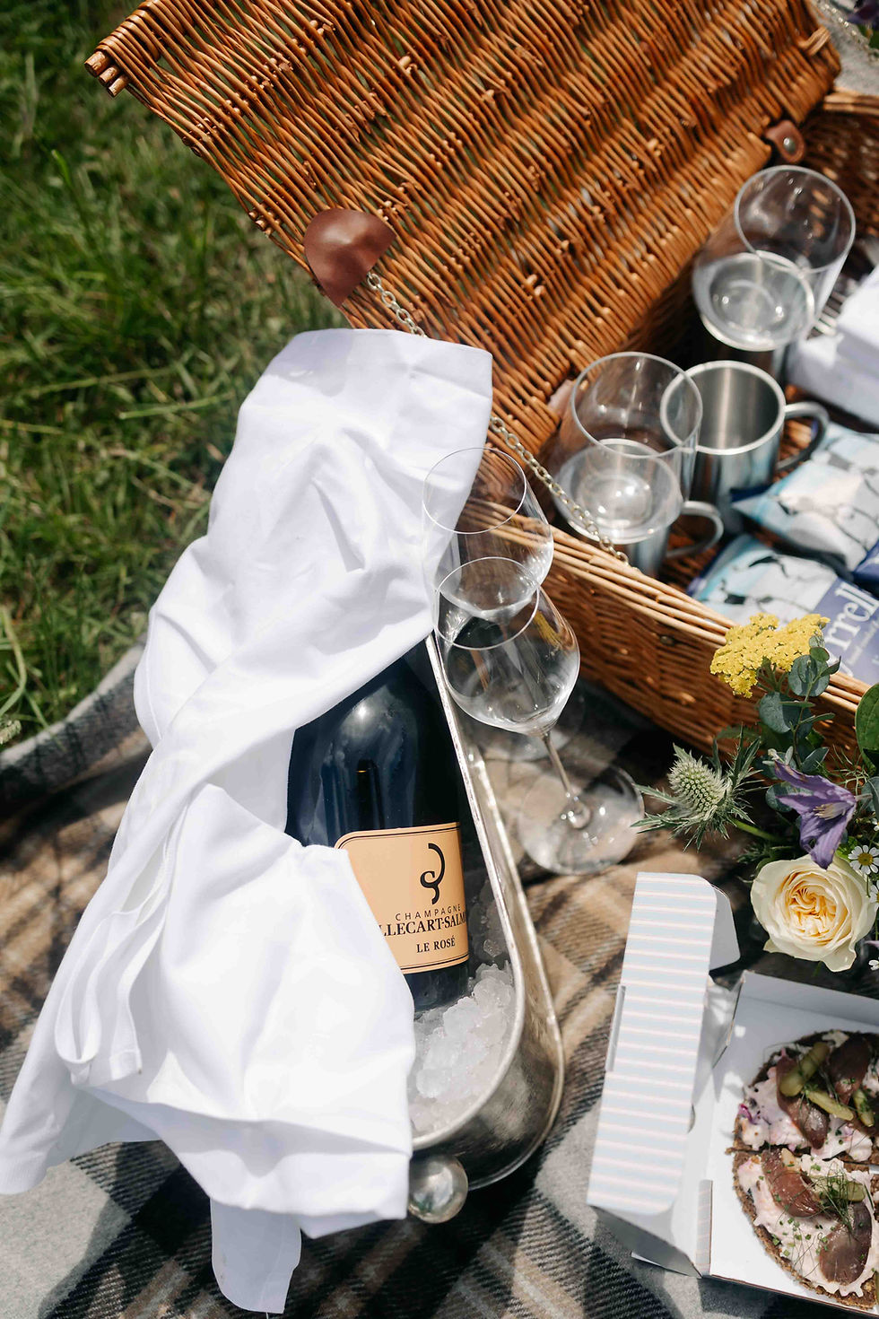 Outdoor picnic basket with Champagne Le Rosé, wine glasses, white cloth, flowers, and plates on a blanket in the grass.