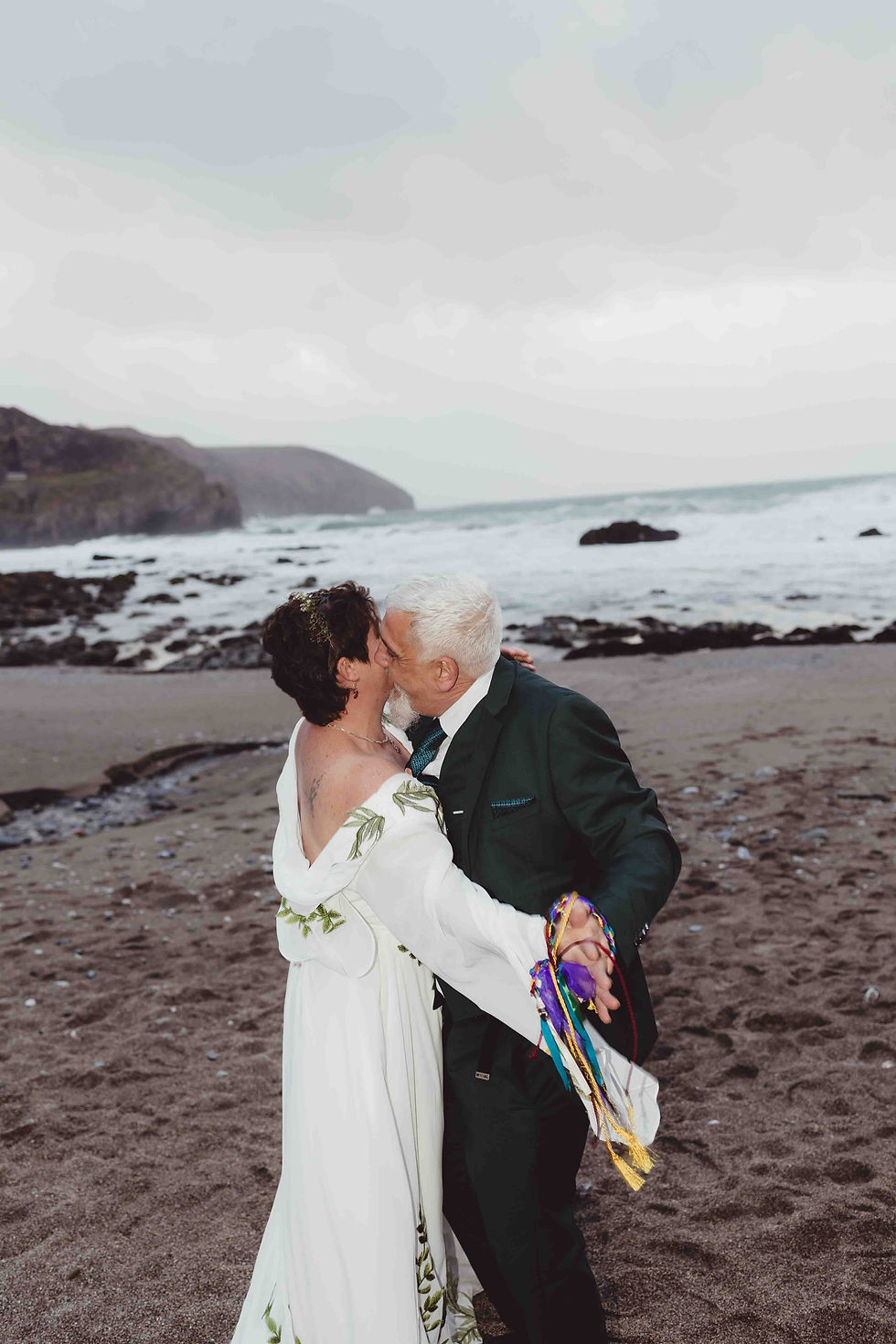 A couple kisses on a rocky beach. They're holding hands tied with colorful ribbons. The sky is overcast, and waves crash in the background.