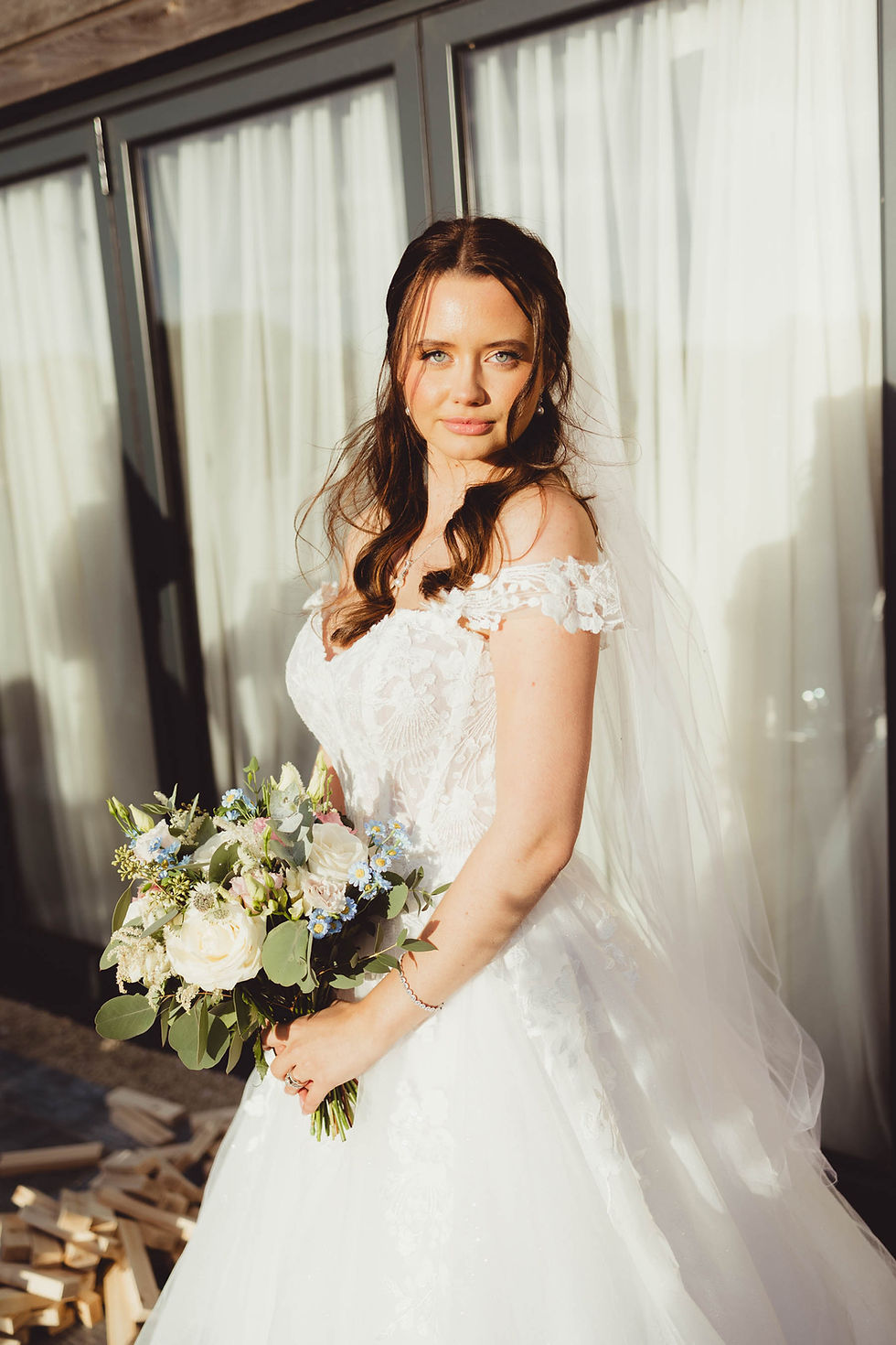Bride in a white lace gown holds a bouquet of white and blue flowers. She stands outside with sunlight on her, beside glass doors.
