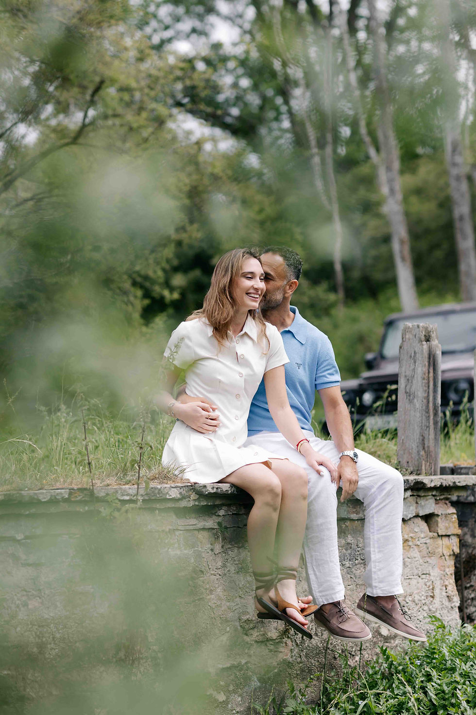 Smiling couple sits on a stone wall in a green park, the man kissing her cheek; she wears a white dress, he a blue polo.