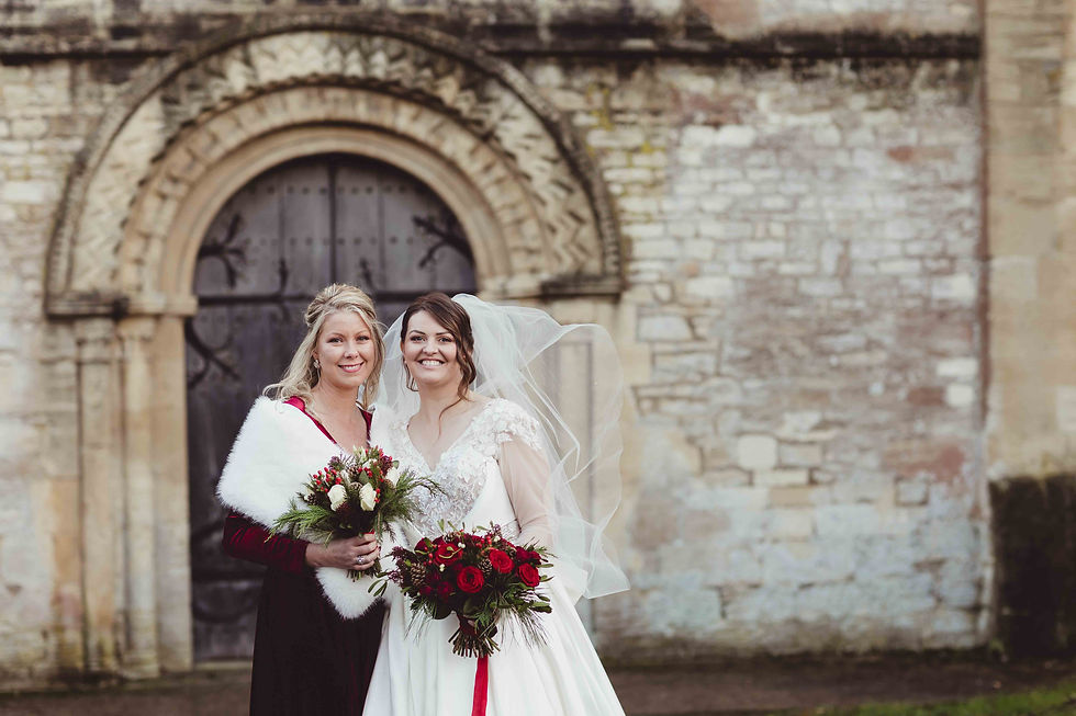 Two smiling women in wedding attire holding bouquets. They stand in front of an ornate stone archway. The mood is joyful and elegant.