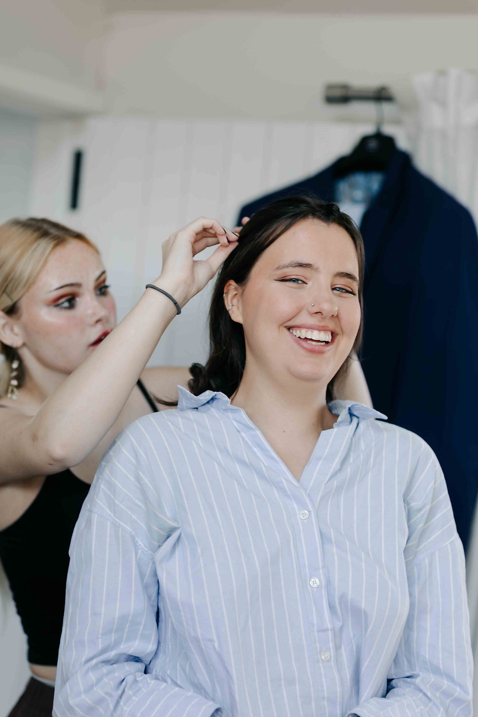 Smiling woman gets her hair styled by another woman in a bright room, with a dark blue jacket hanging in the background
