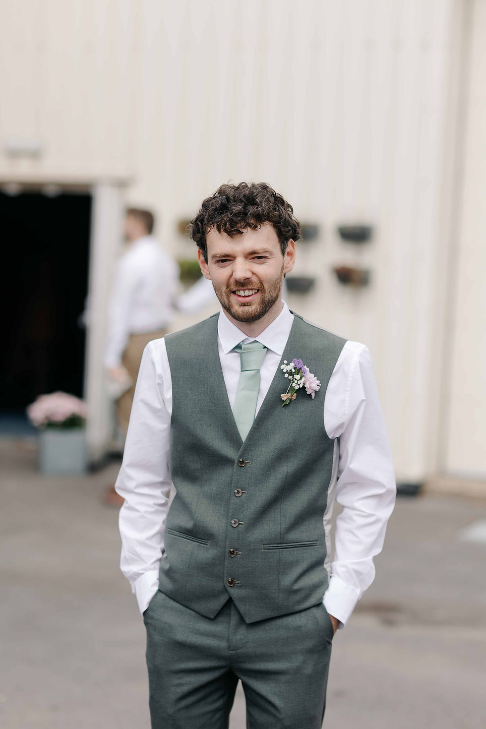 Smiling groom in a sage green vest and tie with boutonniere, standing outdoors at a wedding venue with blurred guests behind him.