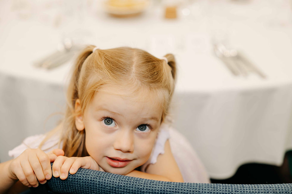 Young blonde girl with pigtails leans on a blue chair, looking up at the camera with a curious expression.