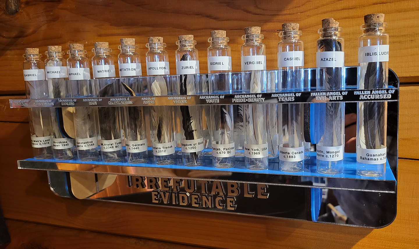 Museum-style specimen display with twelve test tubes holding feathers, each accompanied by typed labels detailing angel names and historical discovery dates, arranged on a reflective mirrored base.