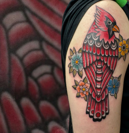 Red cardinal tattoo in traditional style with blue and orange flowers, placed on the upper arm.